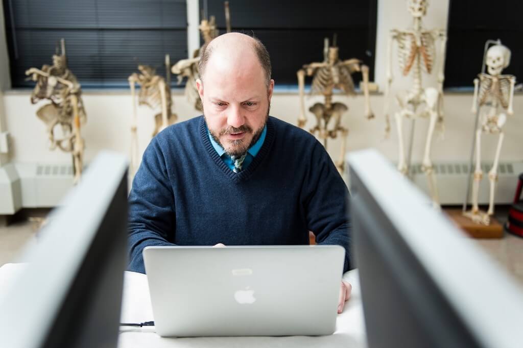 As skeletons hang out close by, UW paleoanthropologist John Hawks types on a laptop in the Biological Anthropology Lab in the Sewell Social Sciences building.